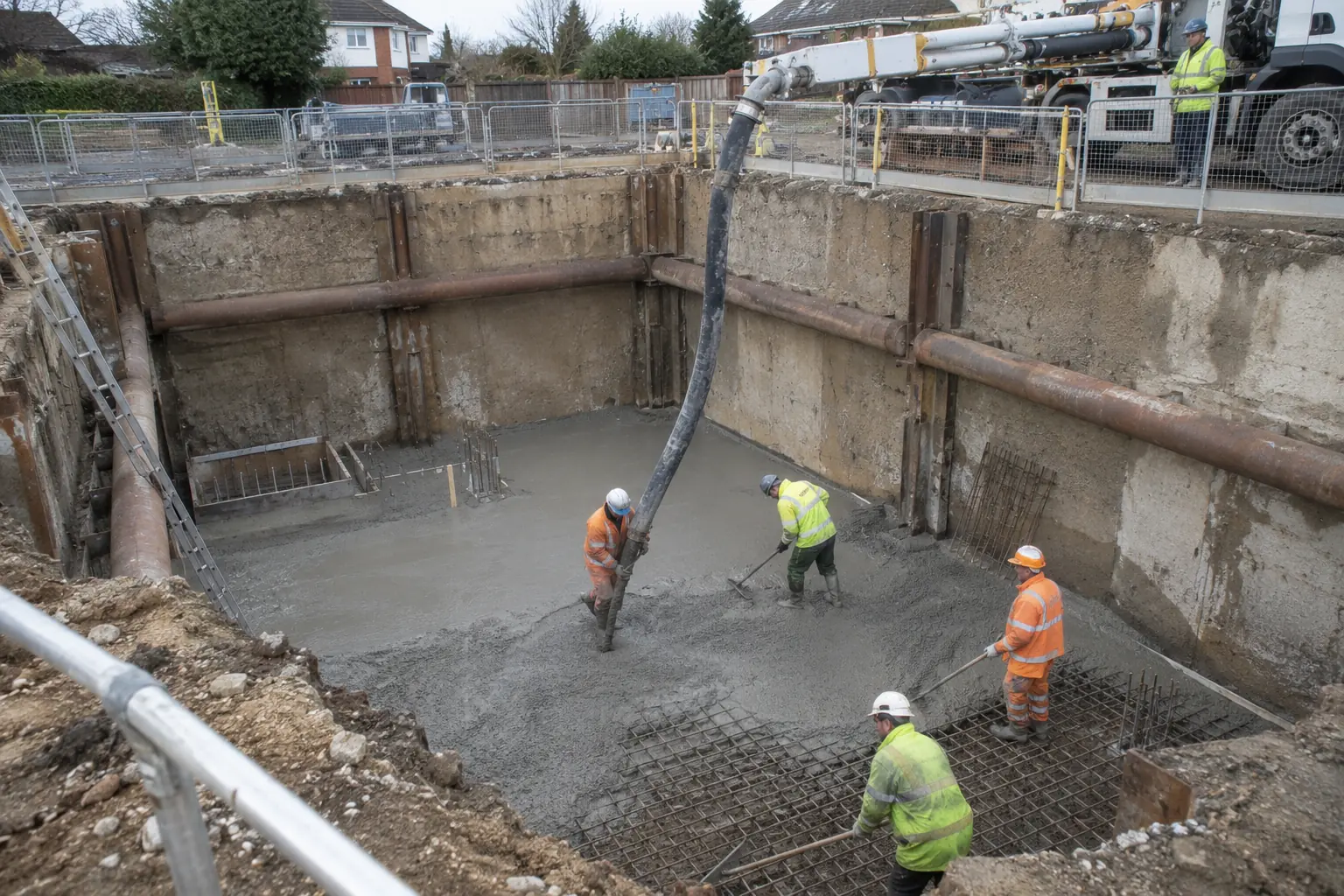 Basement concrete pumping — Wide view down into an open, safely shored basement excavation where a thick concrete delivery hose descends from ground level to a PPE-clad crew placing concrete onto a prepared basement floor. Guarded excavation rim, retaining supports and clear below-ground depth.